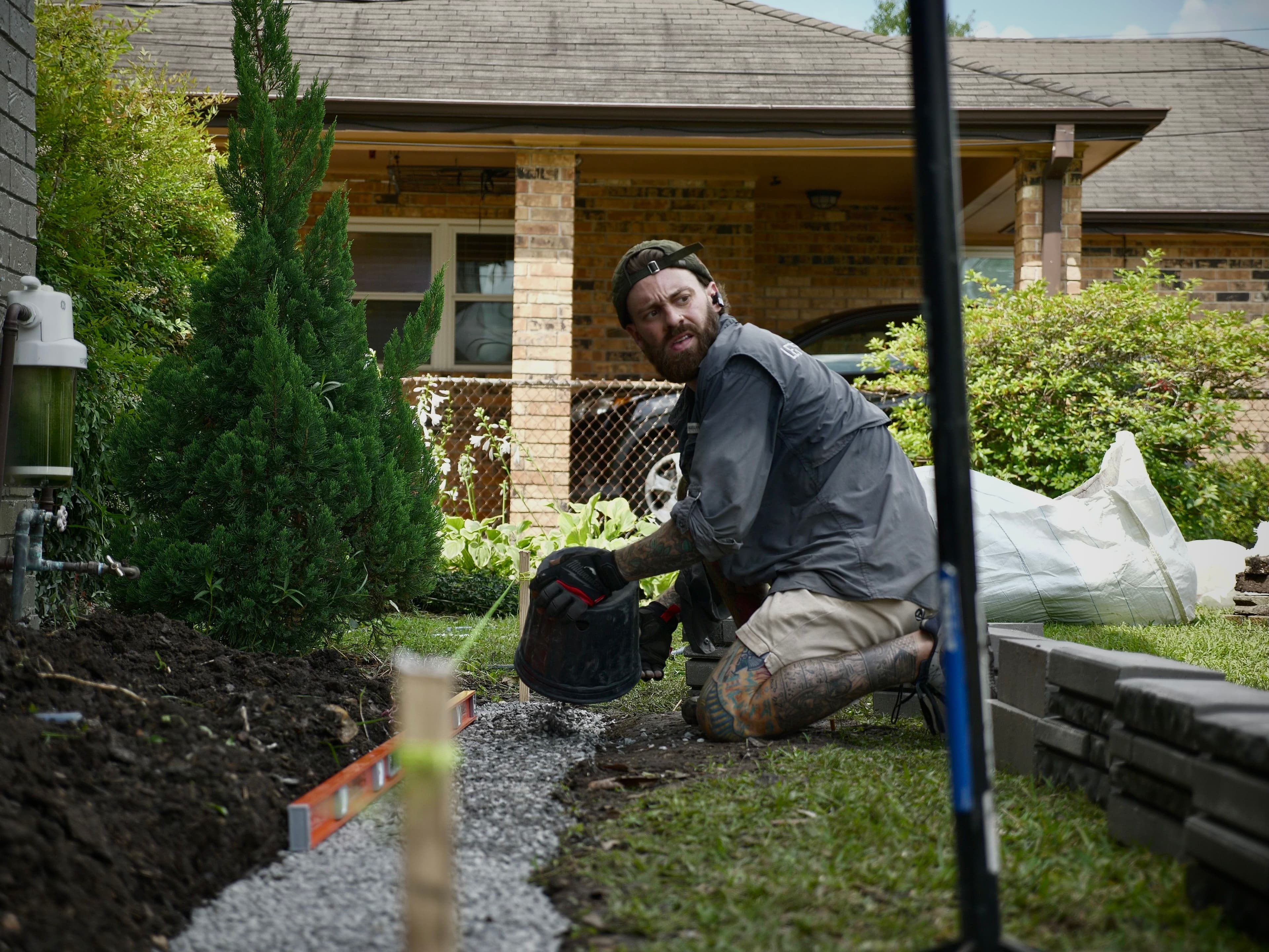 Man landscaping a pathway with gravel in a residential yard, wearing gloves and working focused.
